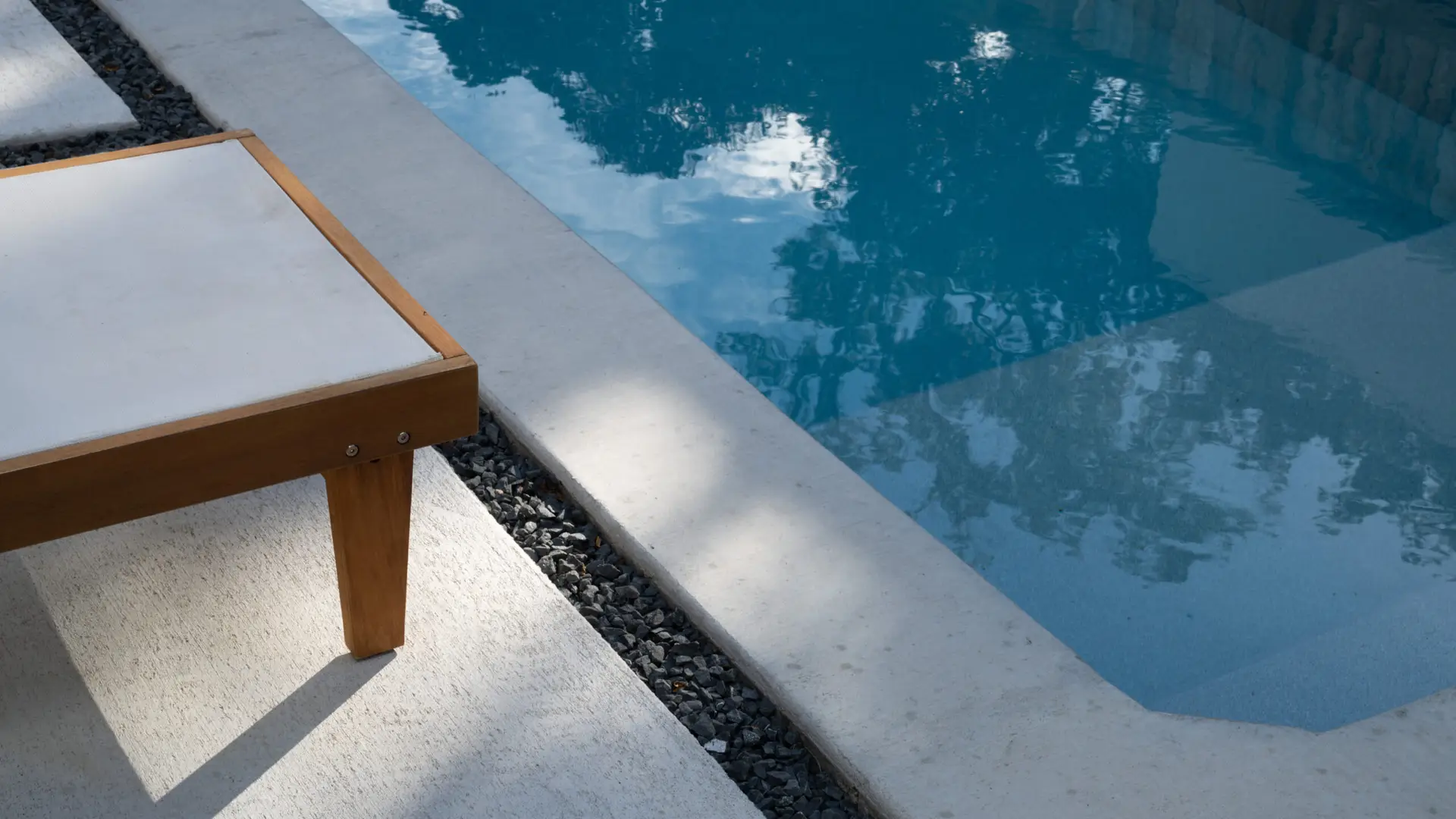 Corner of a modern outdoor table beside a swimming pool, with water reflecting sunlight.